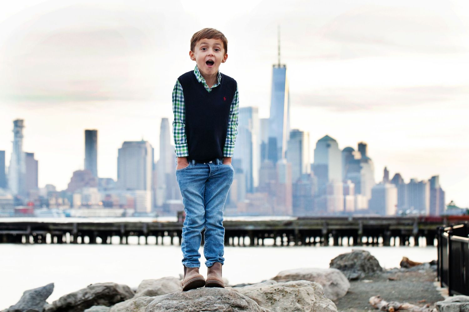 Child standing on rocky waterfront with New York City skyline in background.