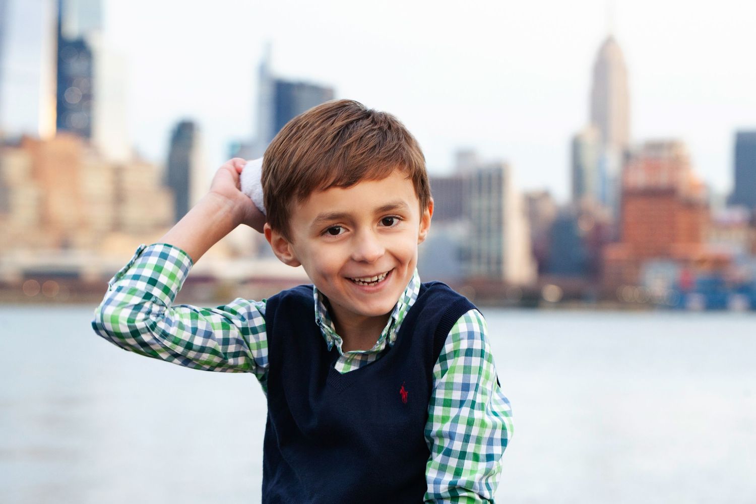 Young child in navy vest and checkered shirt smiles with NYC skyline behind.