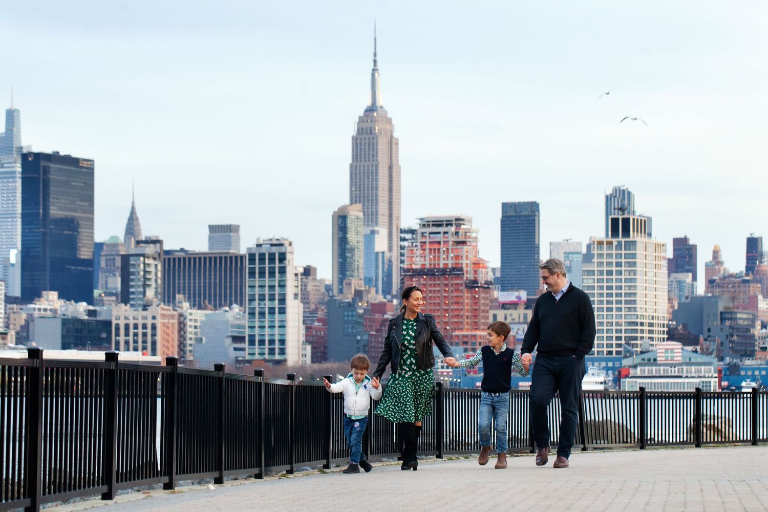 Family walking along waterfront path with Empire State Building in background.