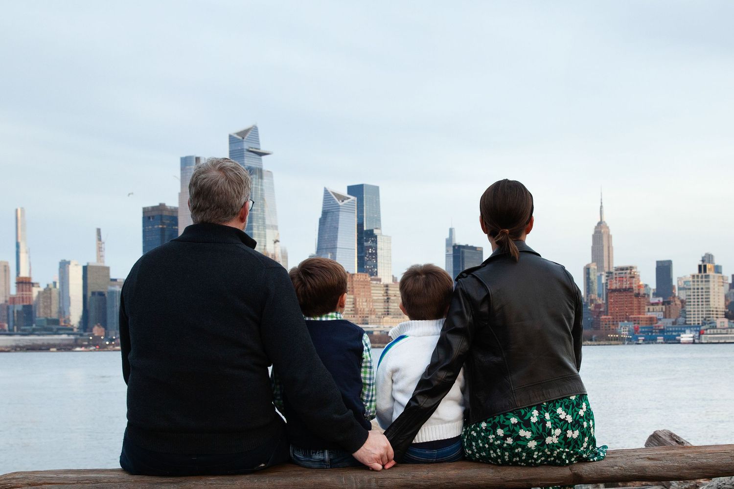 Family sitting together on waterfront viewing New York City skyline at sunset.
