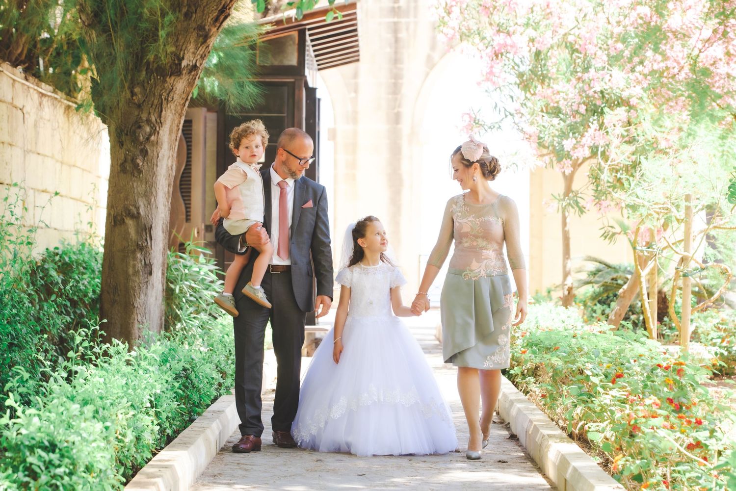 An outdoor wedding scene with people gathered under a covered walkway surrounded by greenery.