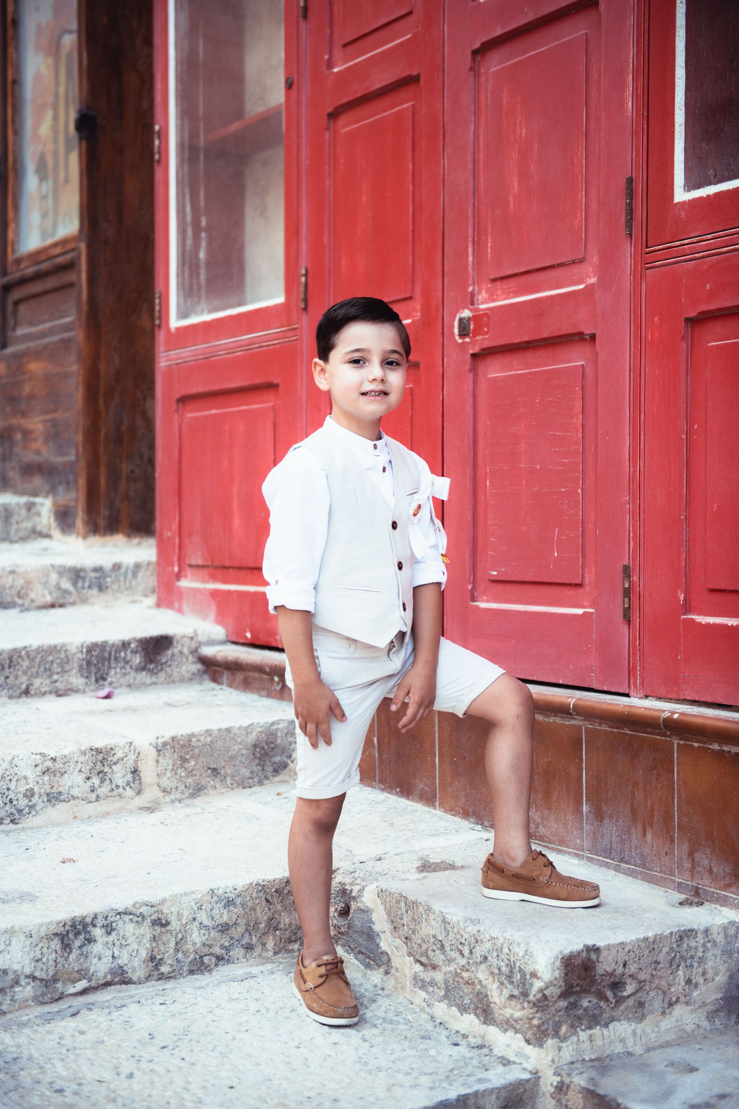 A child in white clothing sits on red painted stone steps in front of a series of red doors.