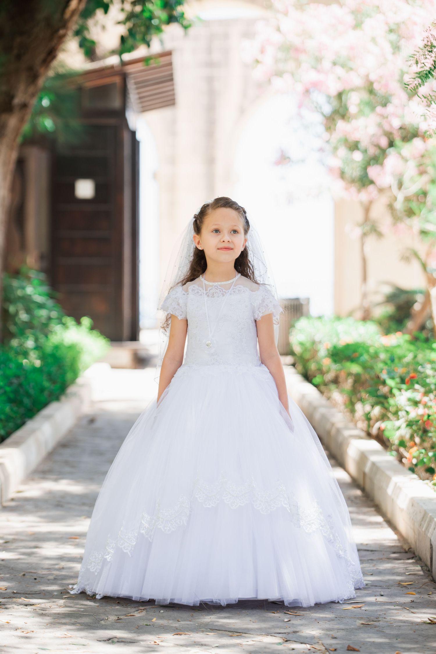 A white lace ballgown photographed in a garden setting with flowering trees.