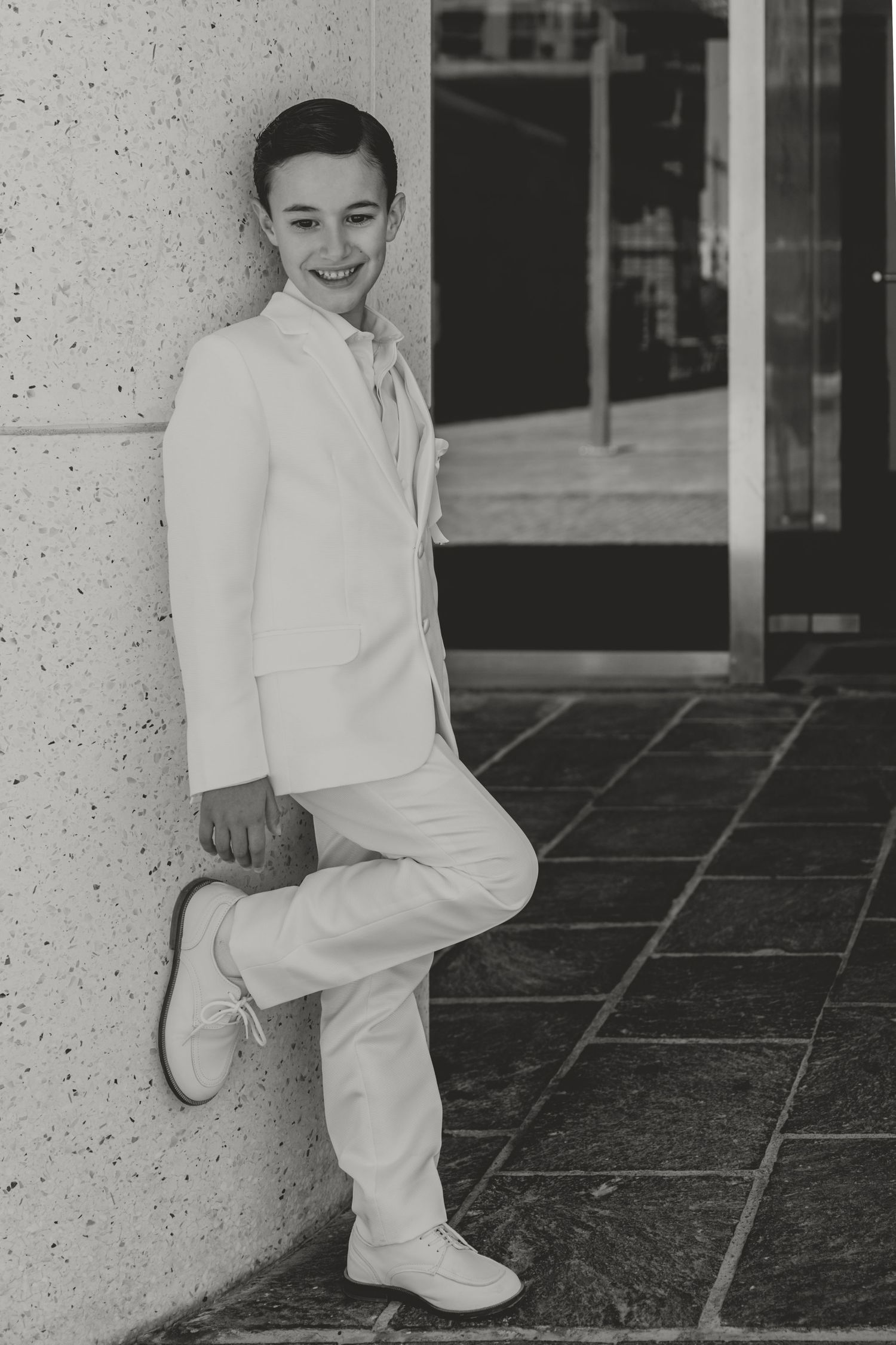 A young person in an all-white suit poses confidently against a wall.