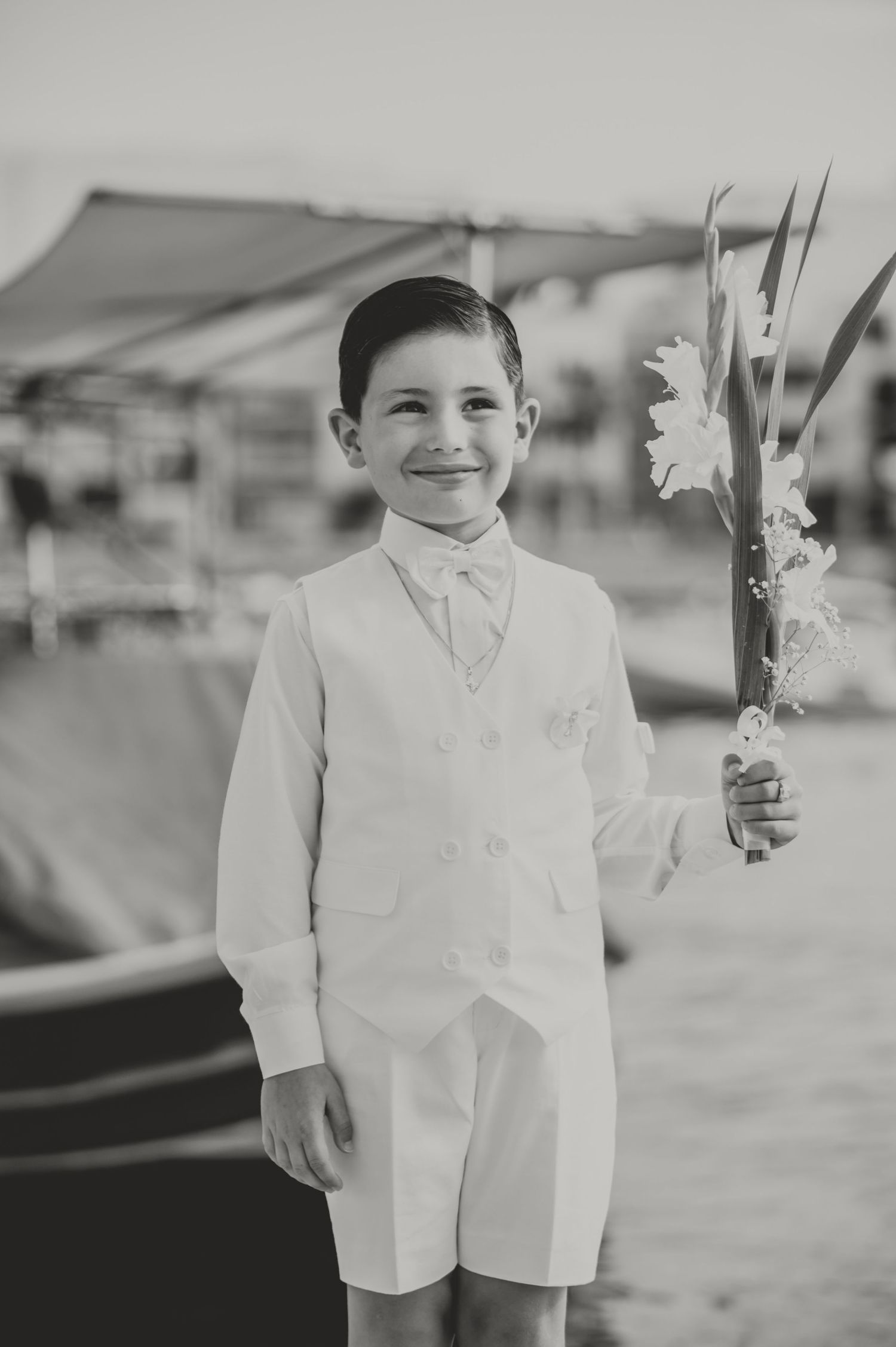A young person in formal white attire holds flowers at a waterfront location.