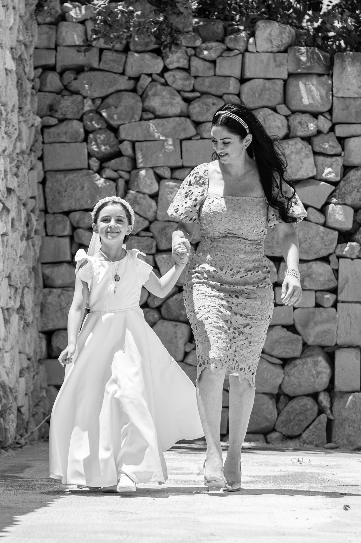 Children in white dresses play and dance in a stone-walled courtyard during a sunny day.