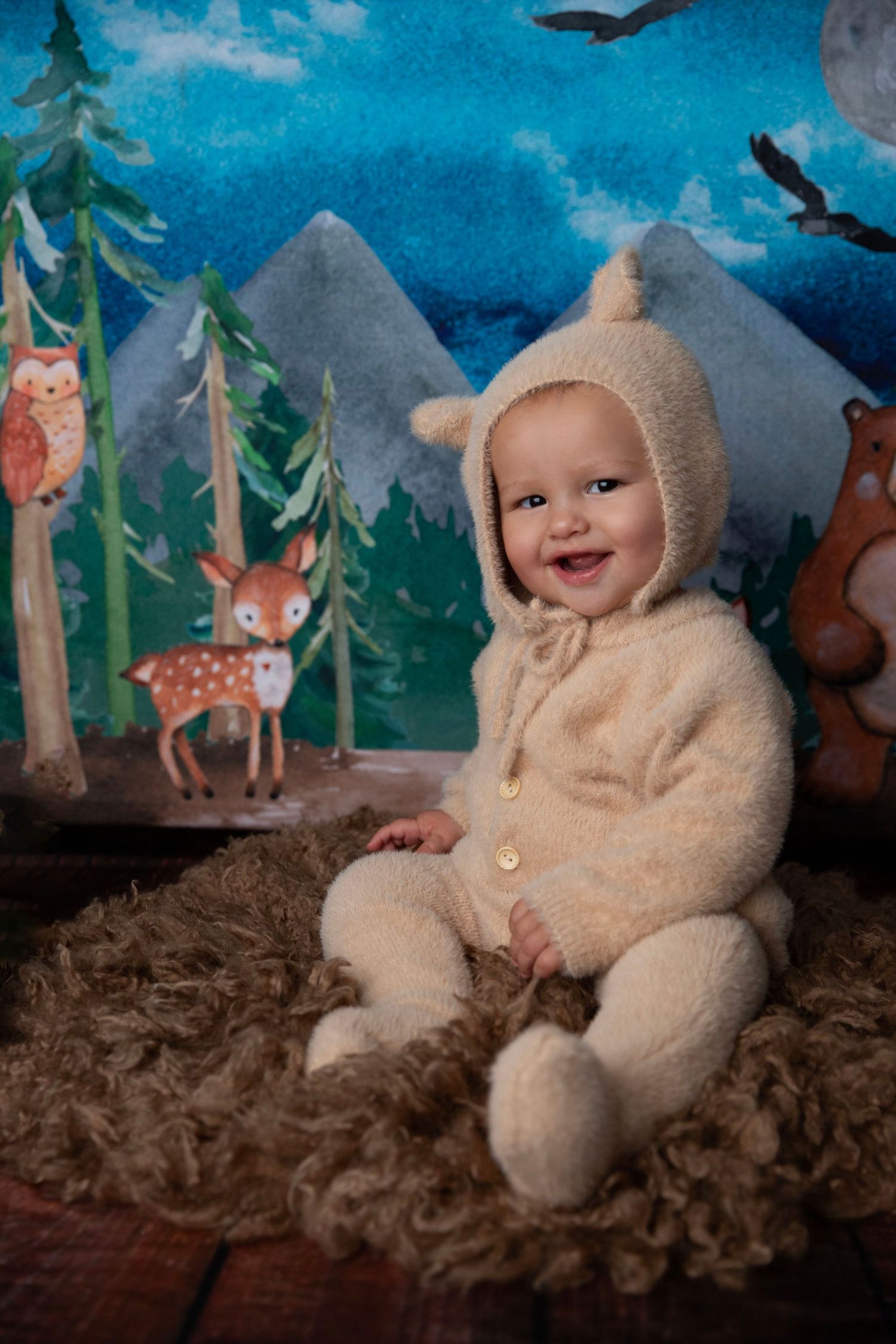 Happy baby in cream fuzzy bear costume sitting on fur rug with woodland animals backdrop.