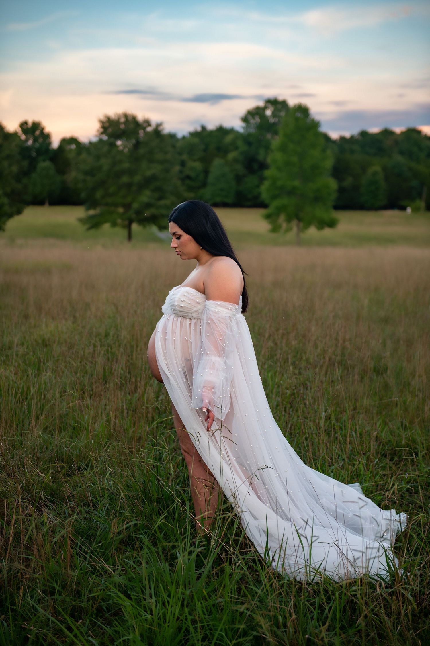 Dreamy maternity session in white lace dress amid tall grass field at dusk.