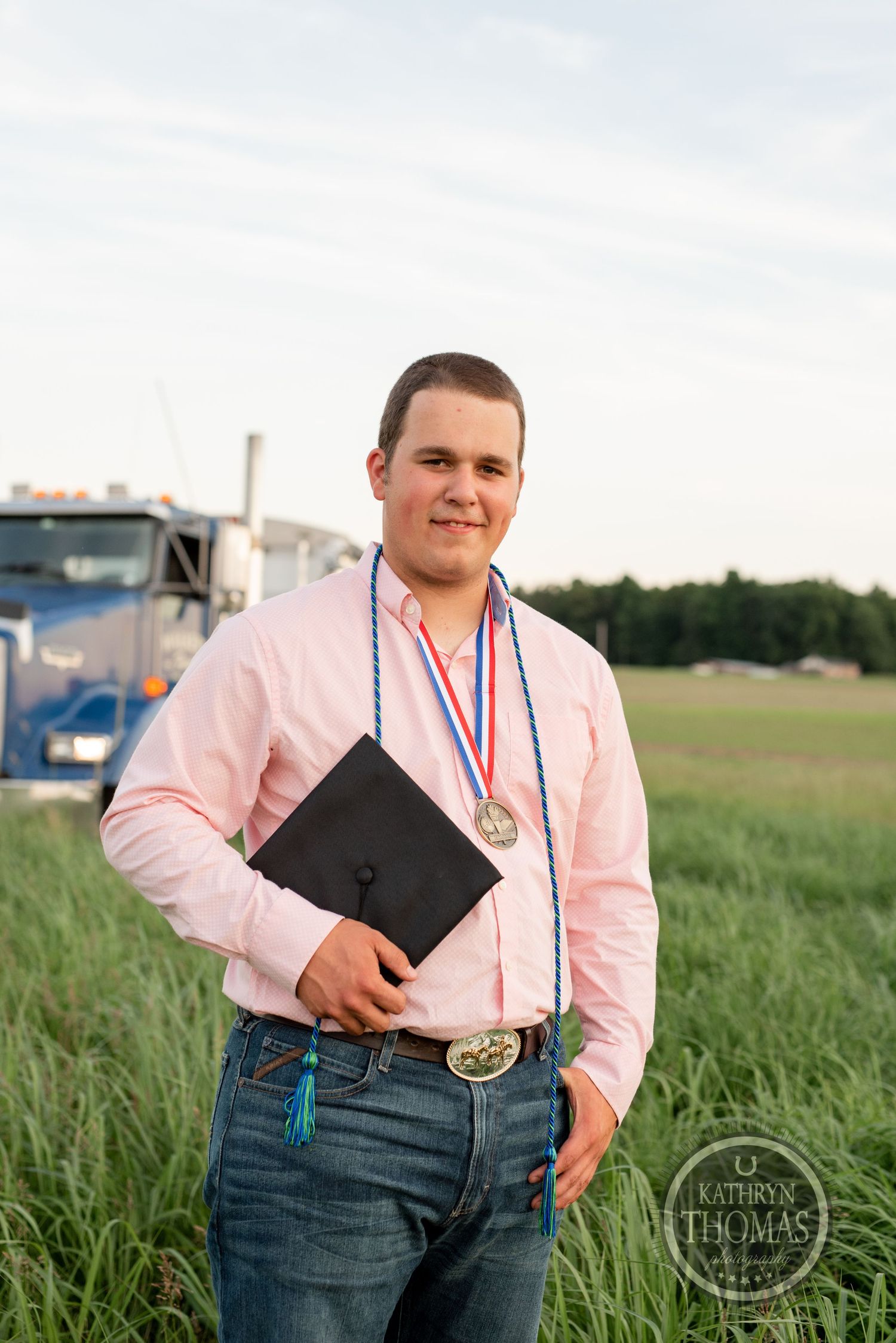 Graduate in pink shirt holding diploma with medal and honor cords poses by truck.