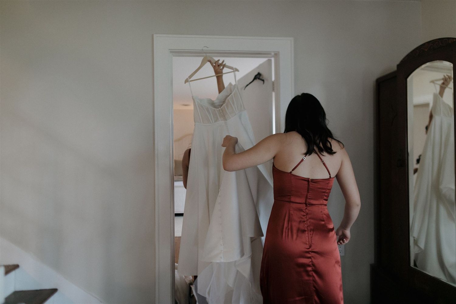 A dress in burgundy helps prepare a white wedding gown hanging in a doorway.