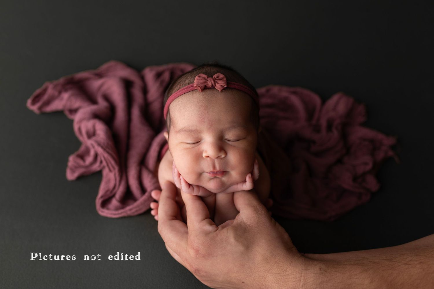 Sleeping newborn cradled in hand against burgundy fabric backdrop.
