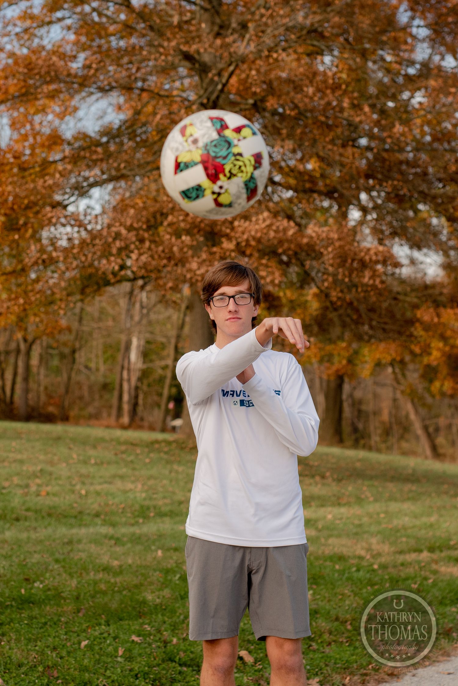 Person in white shirt juggles soccer ball against backdrop of fall foliage.
