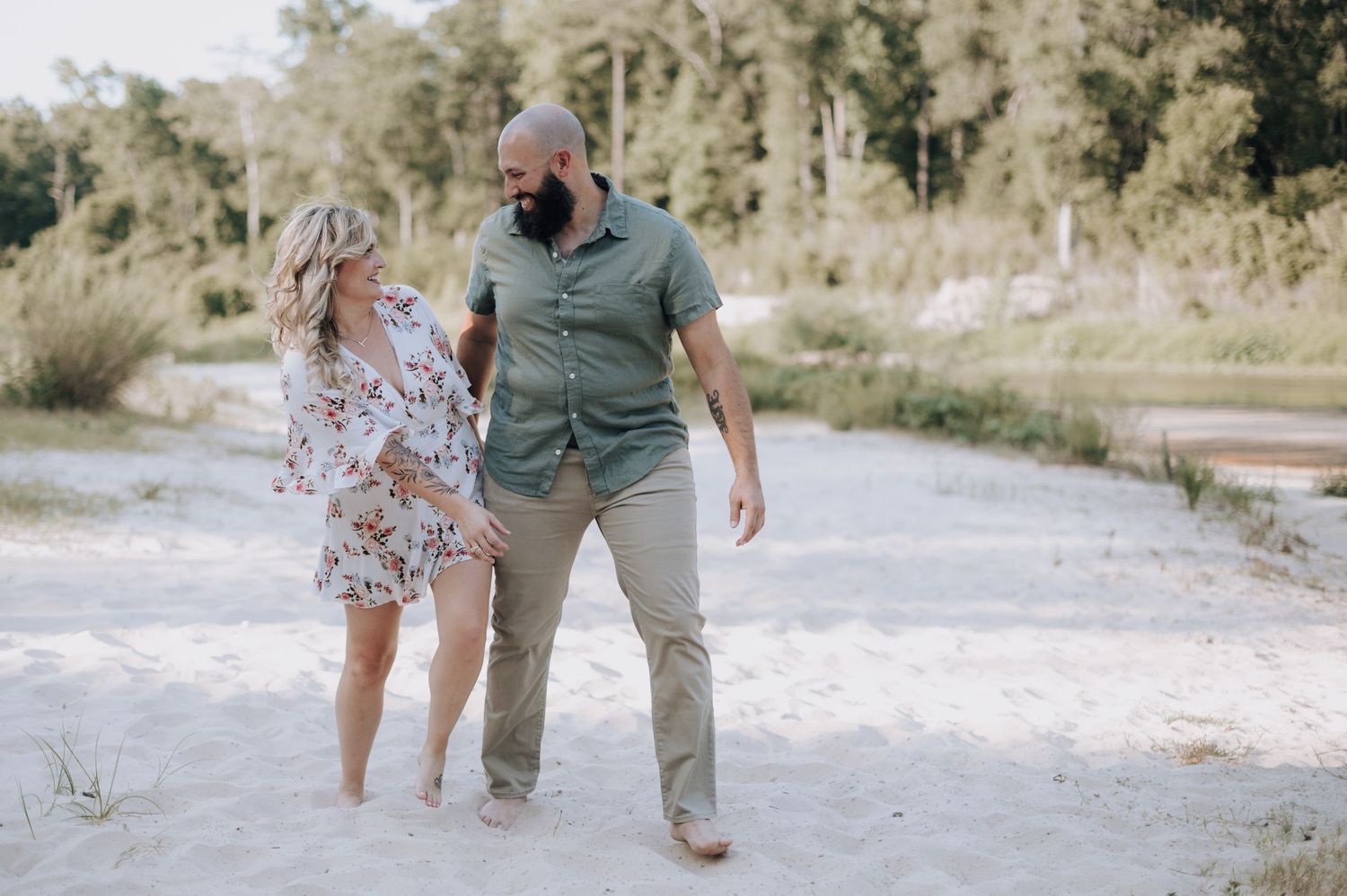 A couple in summer outfits walks together laughing on a sandy trail surrounded by trees.
