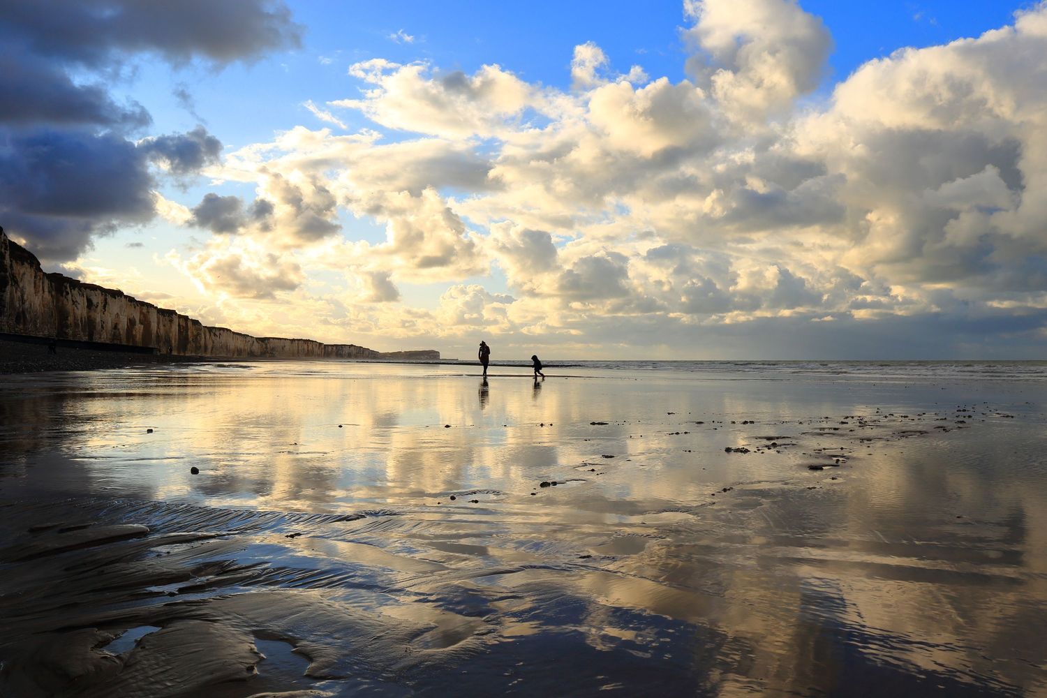 Dramatic sunset sky reflected in wet beach sand with silhouettes in the distance.