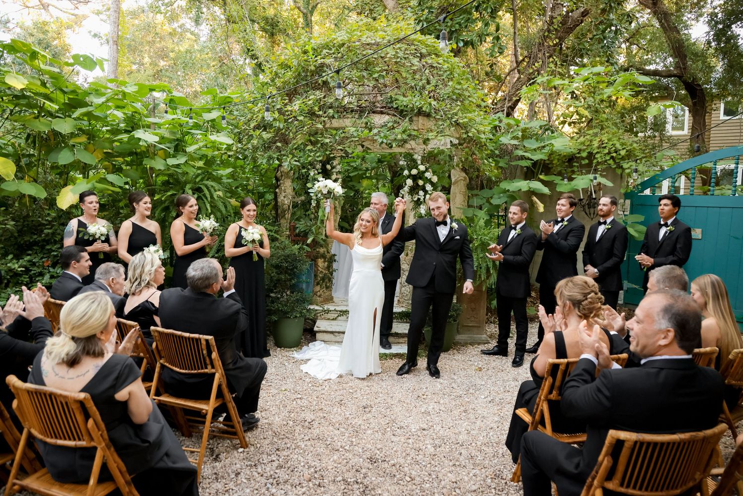 Outdoor garden wedding ceremony takes place on gravel path surrounded by lush greenery and seated guests.