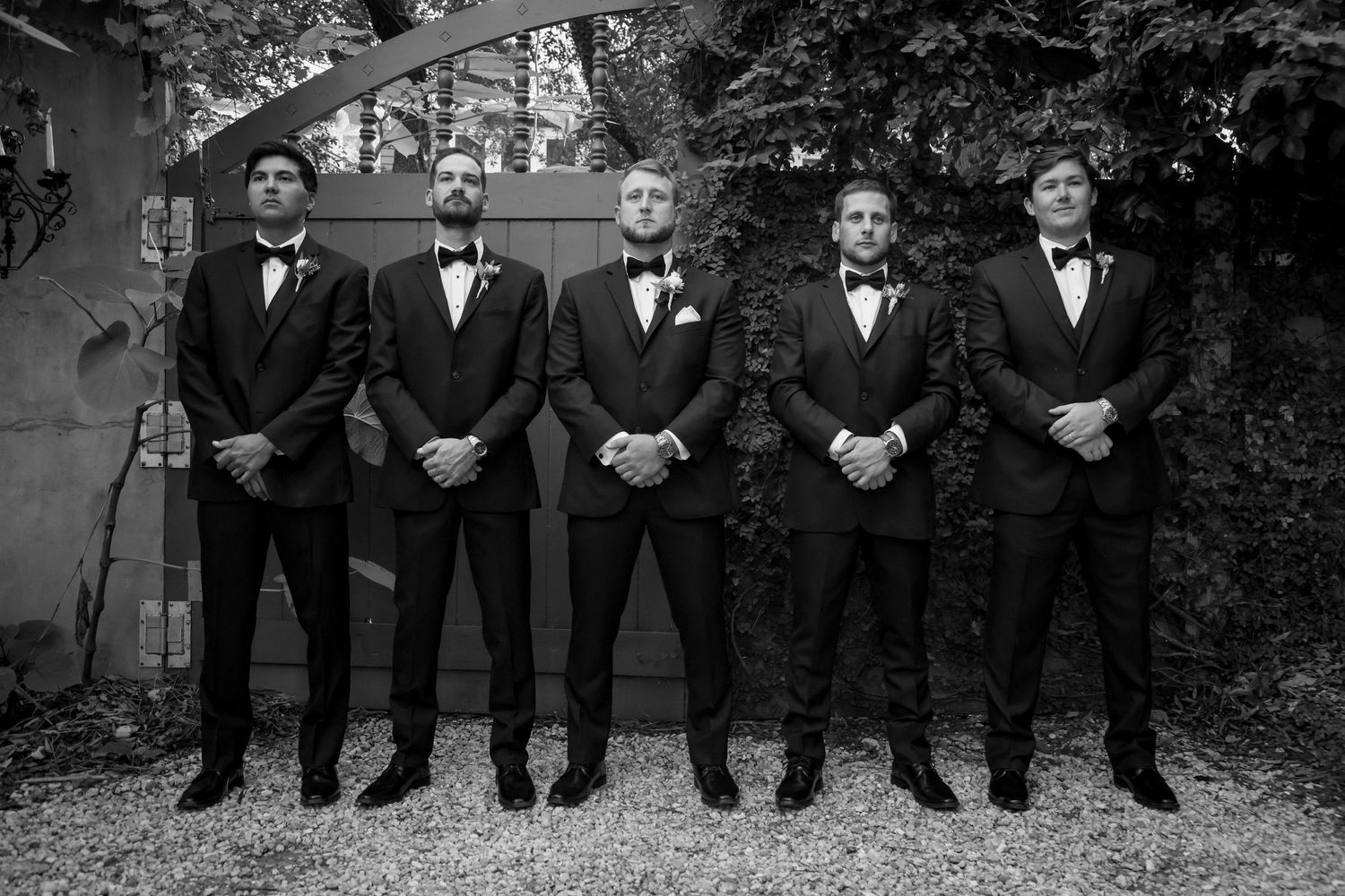 Five groomsmen in classic black tuxedos and bowties stand in a line outdoors for a formal wedding photo.