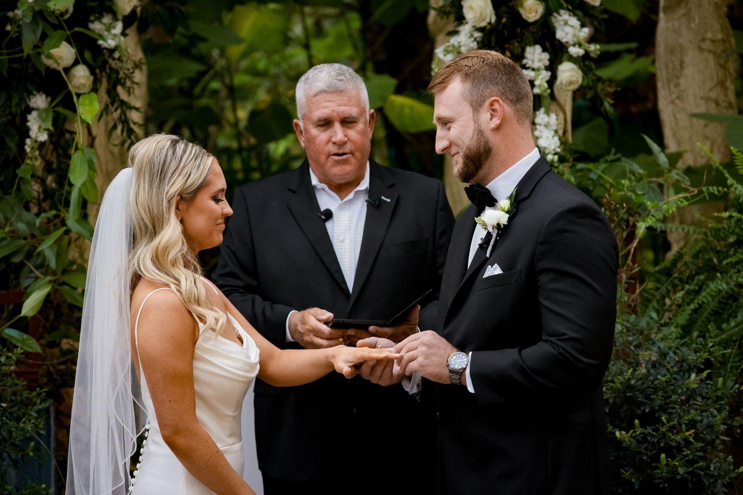 A couple exchanges rings during an outdoor wedding ceremony with an officiant in a lush garden setting.