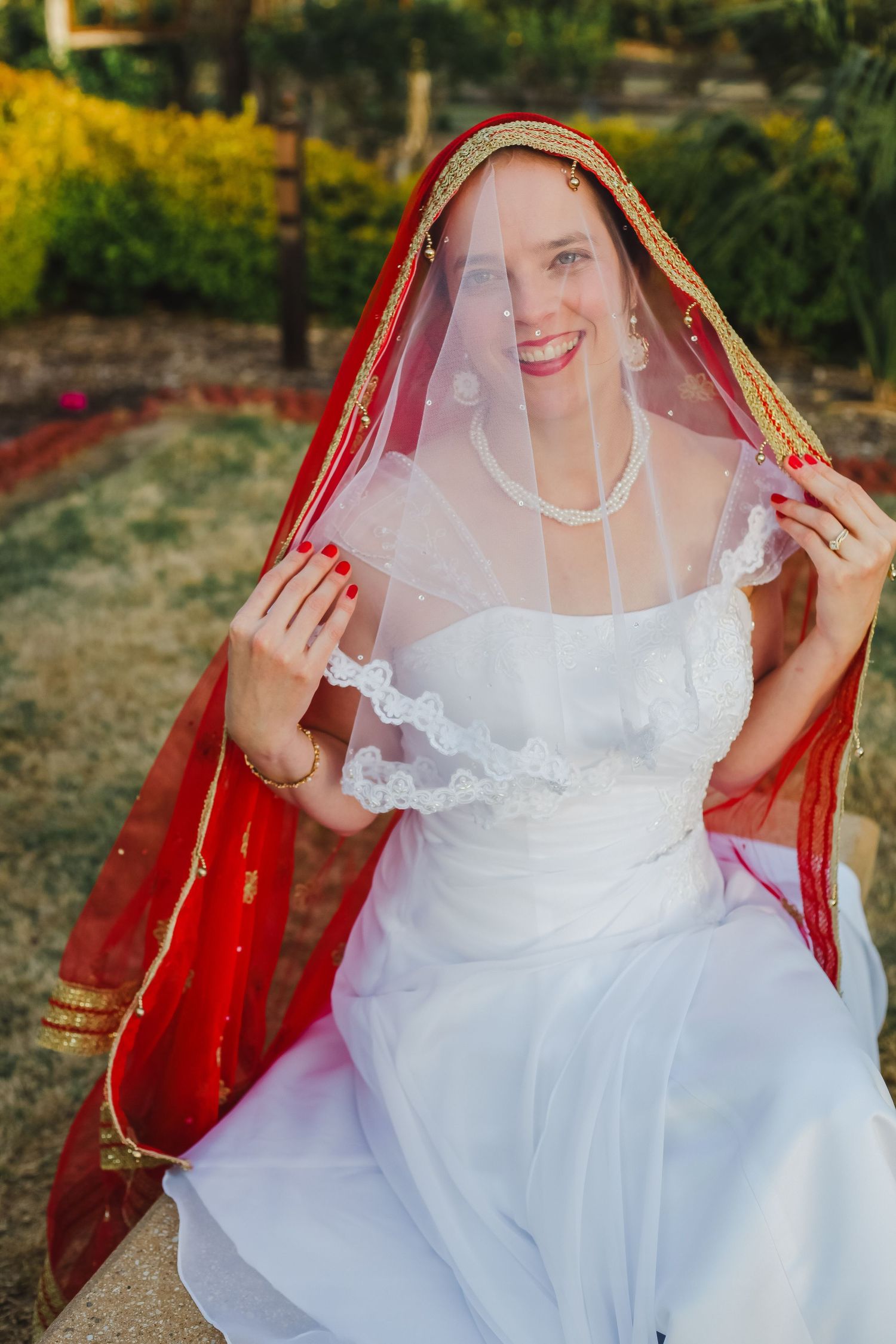 White wedding dress paired with red and gold veil in outdoor garden setting.