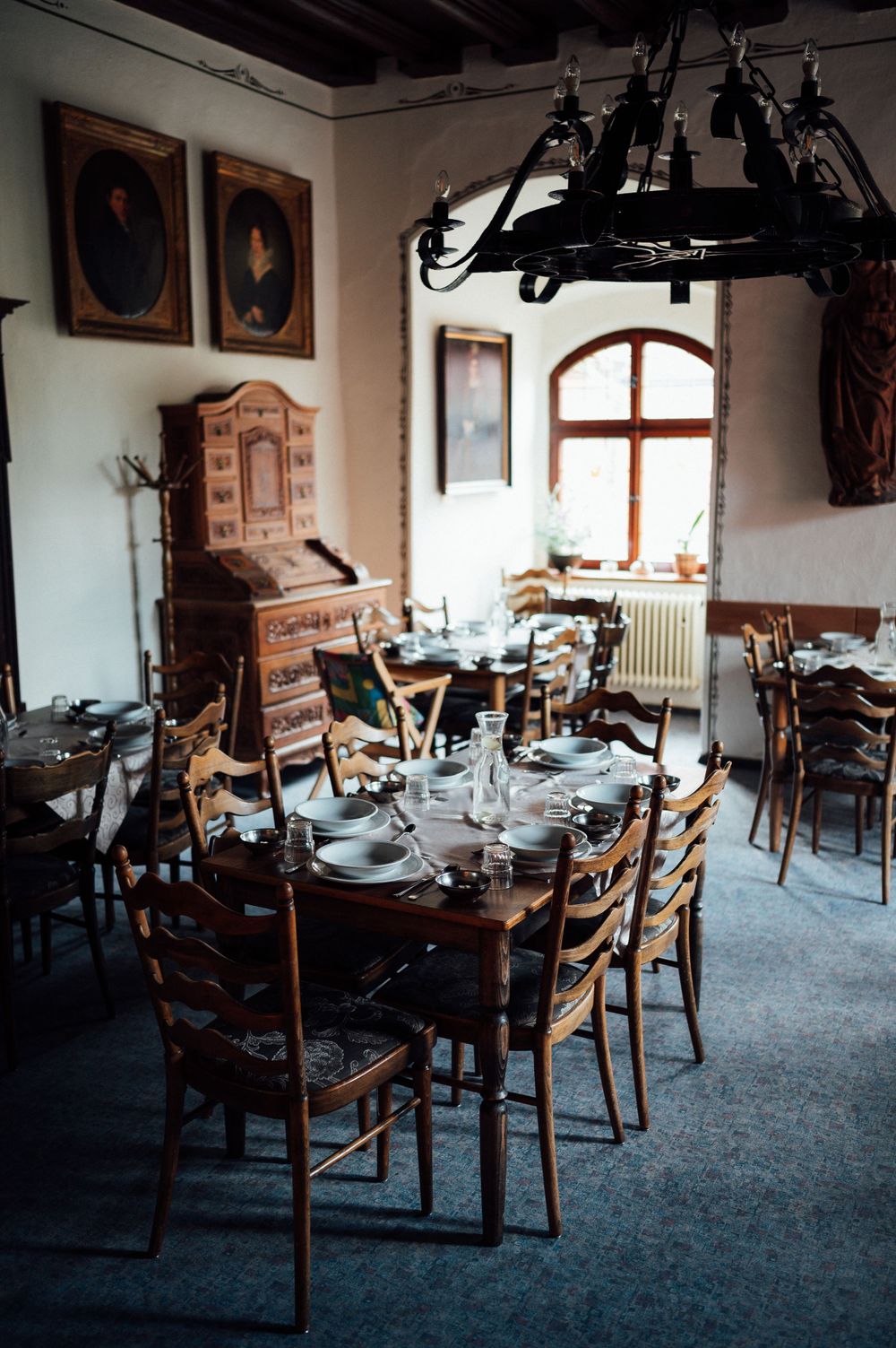 A rustic dining room with antique furniture, portraits on walls, and a wrought iron chandelier.