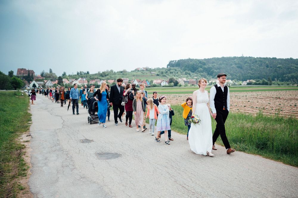 A wedding procession walking along a rural path with fields in the background.