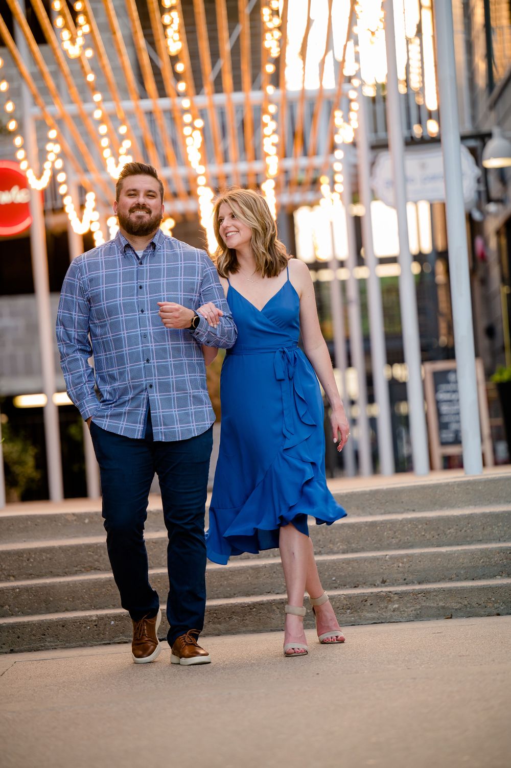 Couple strolls together under string lights, woman wearing flowing blue dress.Congress Avenue Engagement session Austin