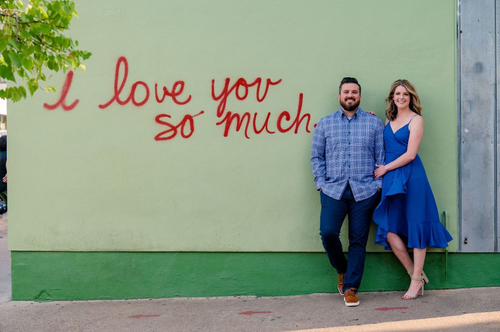 A couple poses against a green wall with red text reading 'I love you so much'. Congress Avenue Engagement session Austin