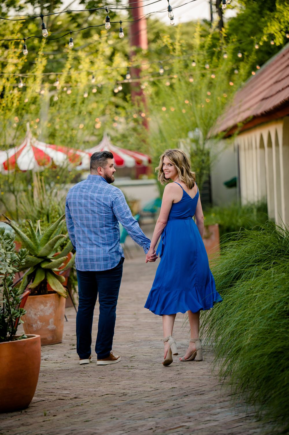 Couple walking hand in hand on garden path with string lights overhead.Congress Avenue Engagement session Austin