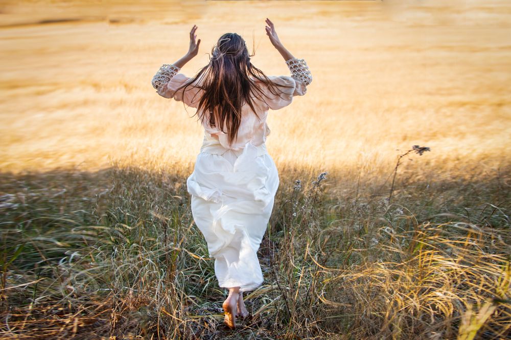Figure in white dress raises arms while standing in golden wheat field at sunset.