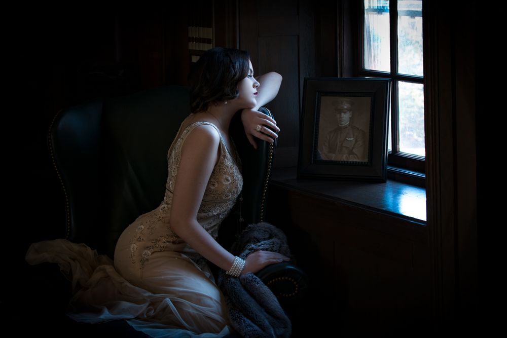 A moody interior scene with someone in vintage dress gazing out a window.
