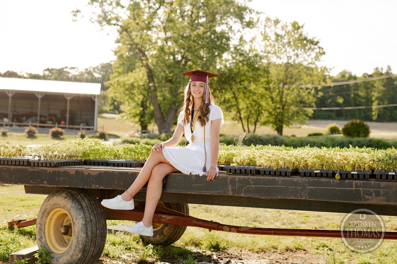 Graduate sits on vintage farm wagon wearing white dress and maroon cap in sunny garden setting.
