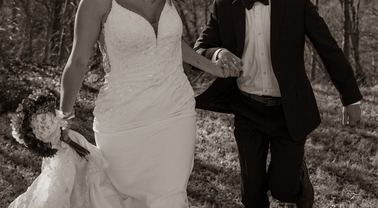 A black and white wedding photo showing a couple walking through a wooded area.