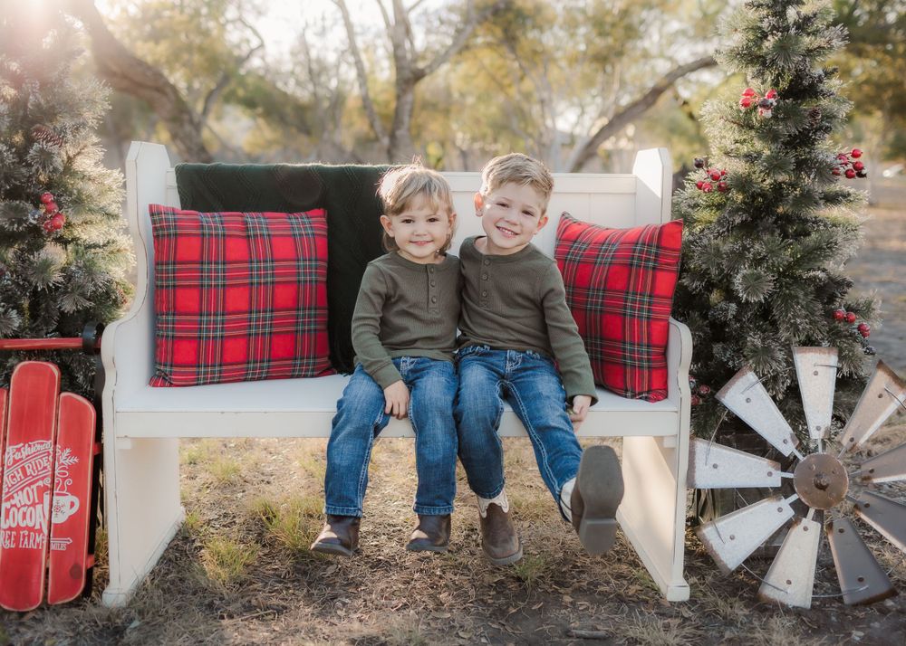 Brothers in green sweaters share a laugh on a white bench with festive red plaid pillows.