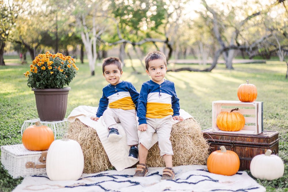 Two children in matching outfits sitting on hay bales surrounded by fall pumpkins.