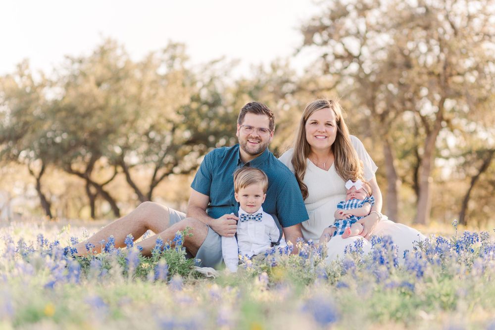 Family sits together in field of bluebonnets during golden hour sunset.