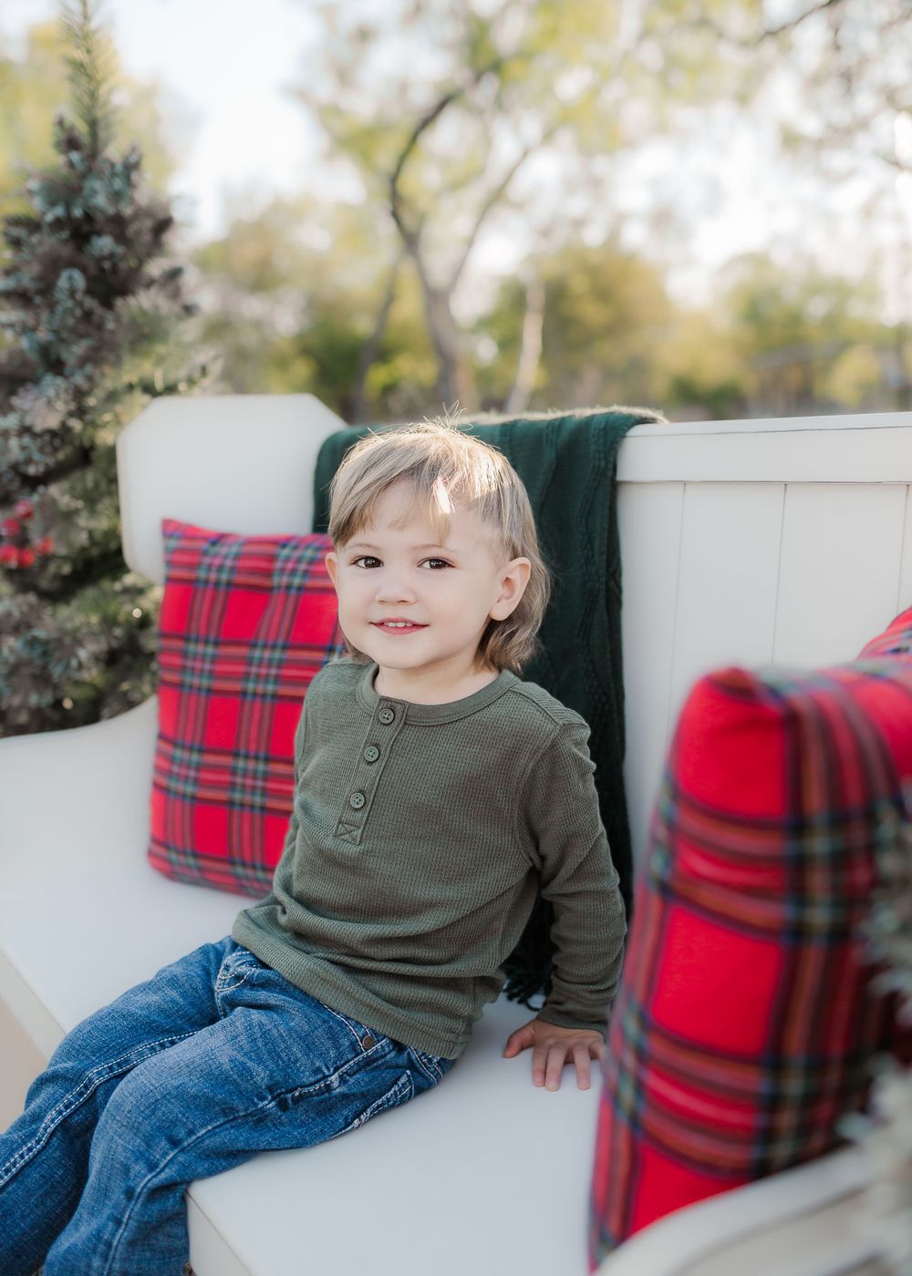 Young boy in green sweater sitting on white bench with red plaid pillows outdoors.
