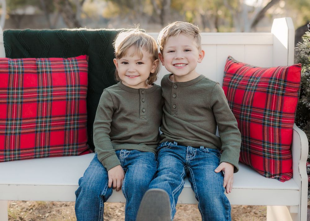 Two young boys in matching sweaters sitting on white bench with red plaid pillows.