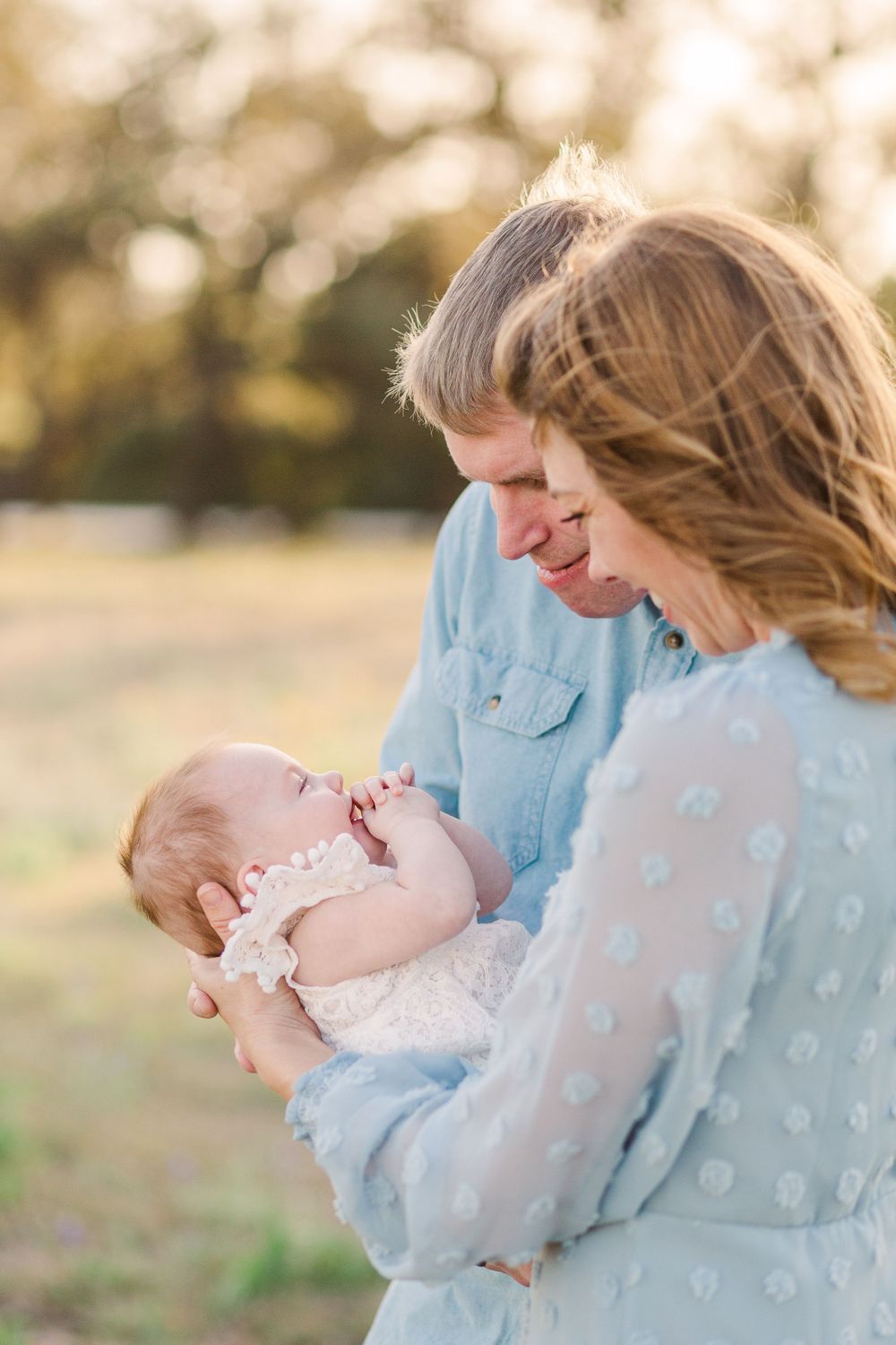 Parents in blue clothing share a tender moment with their baby outdoors in golden sunlight.