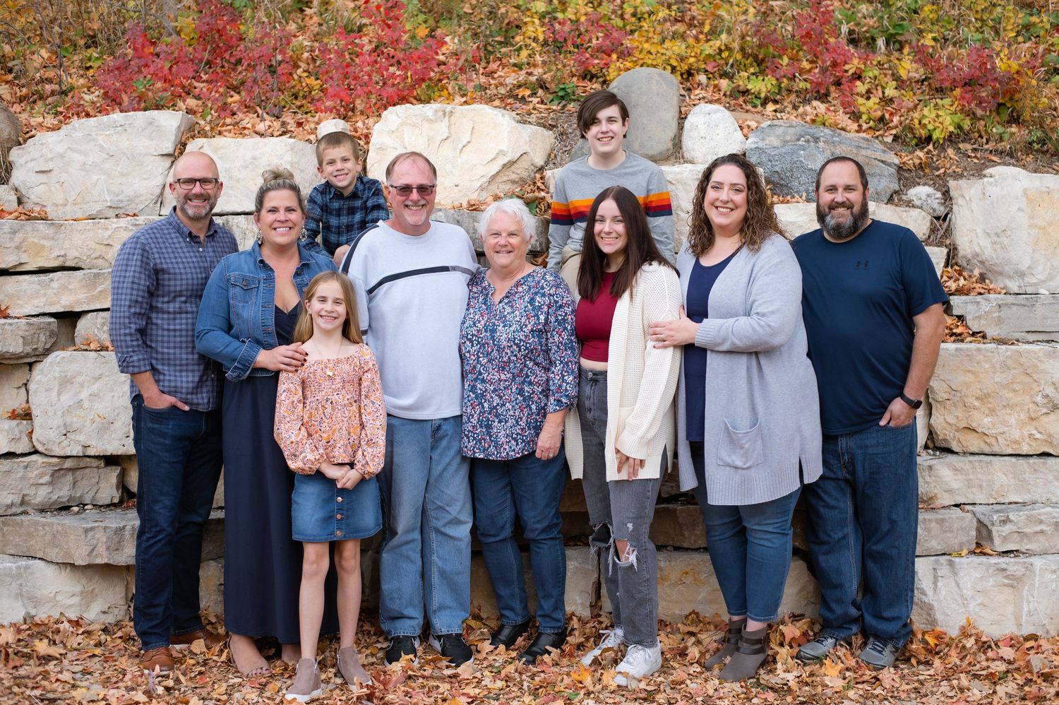 Multi-generational family portrait outdoors against stone wall with fall foliage.