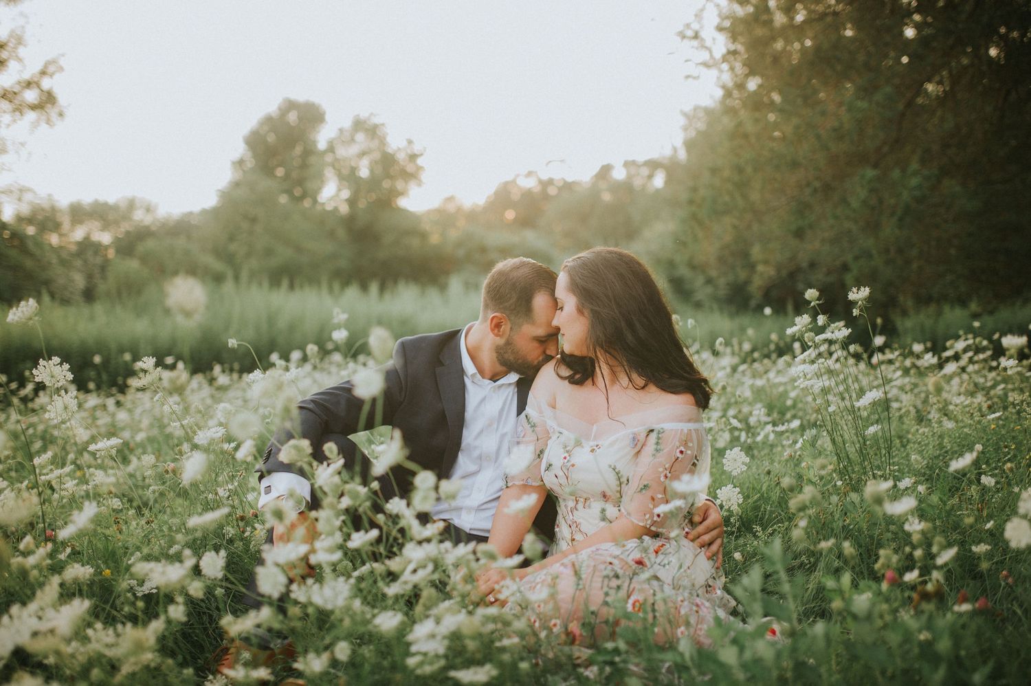 A couple sits together in a wildflower meadow during a romantic engagement photoshoot at sunset.