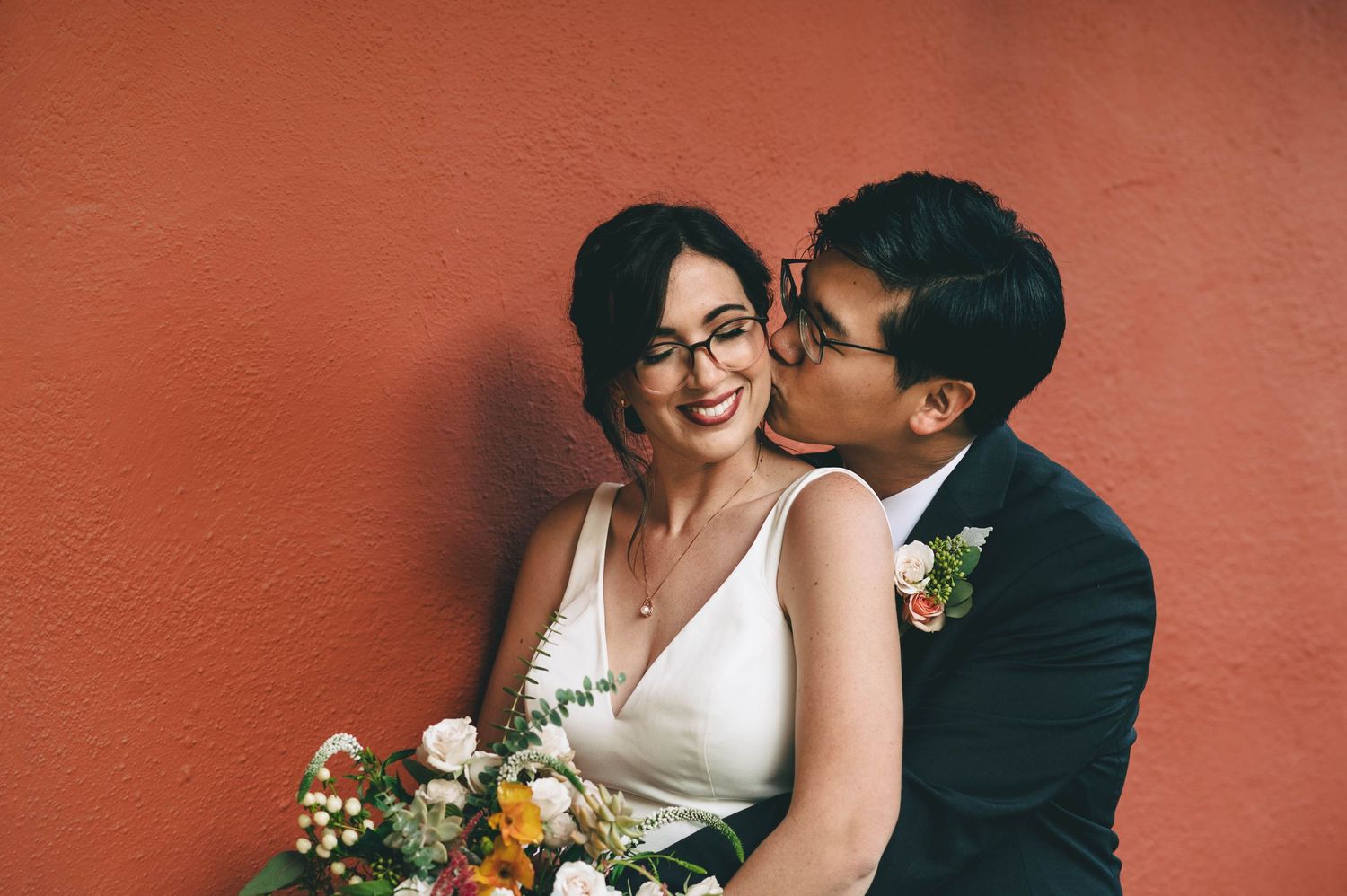 Groom kisses bride on cheek, in front of the red Adobe walls at Cooper Molera in Monterey.