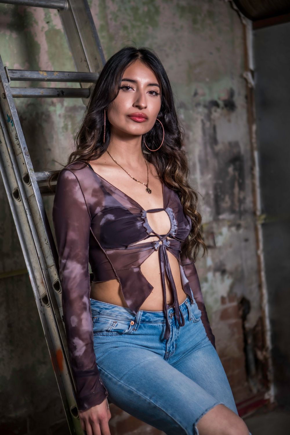 Fashion portrait of model in purple tie-front top and jeans posing against industrial backdrop.