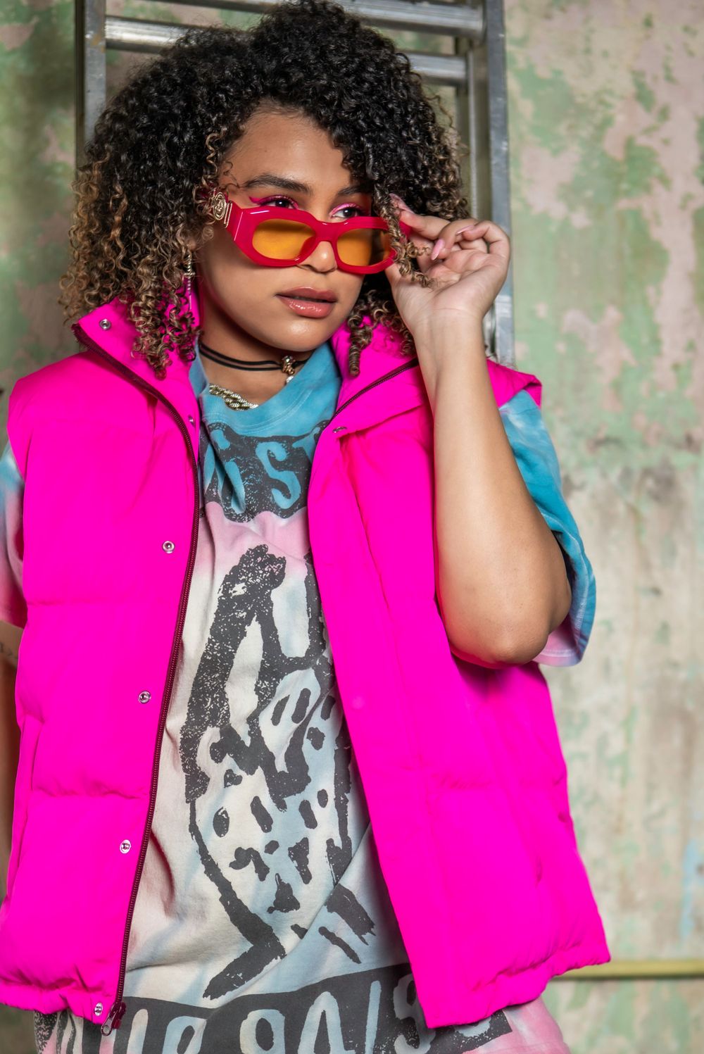 Street style portrait featuring bright pink puffer vest and red sunglasses against weathered wall.