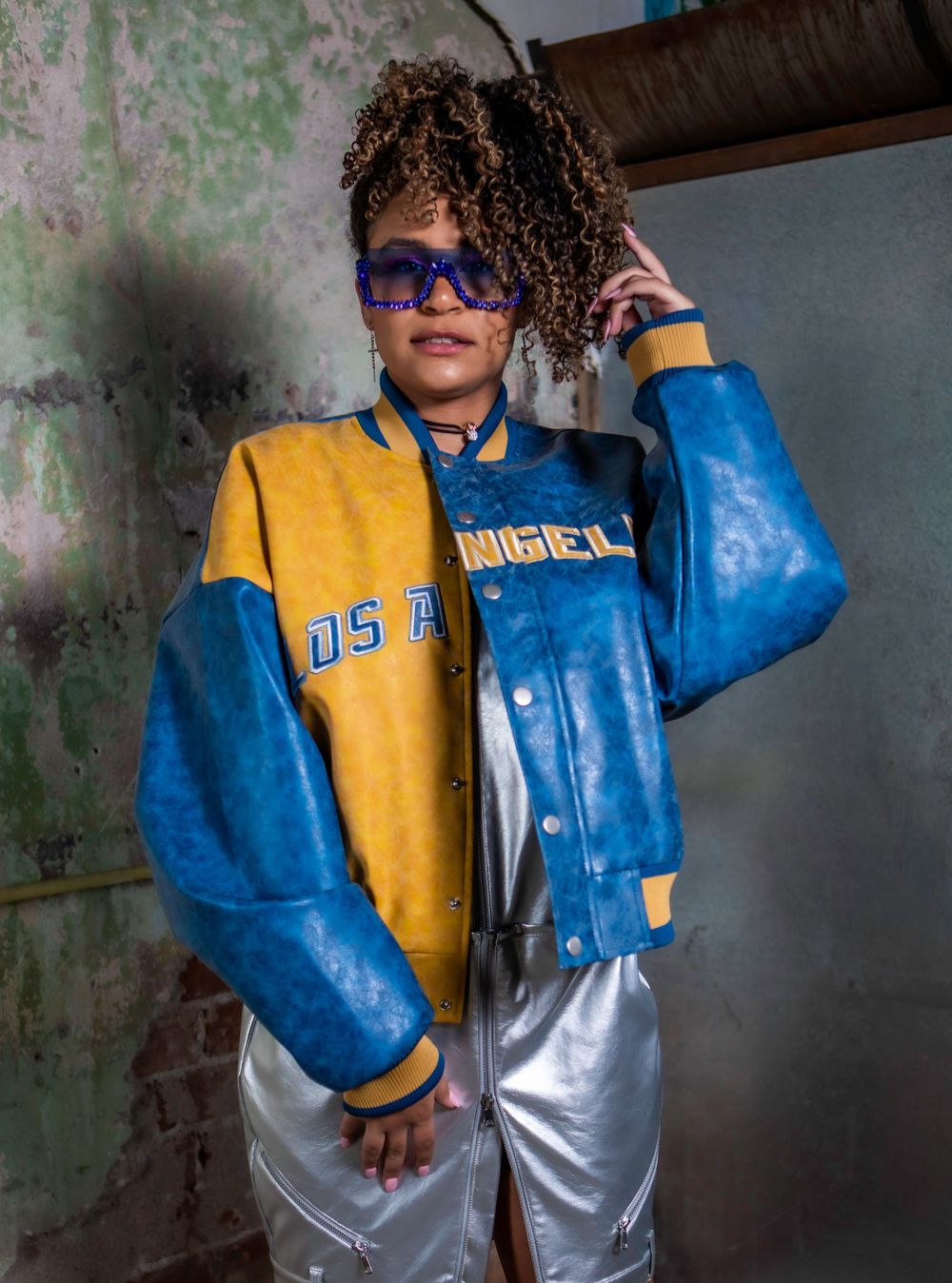 Urban fashion shot featuring blue and yellow varsity jacket against industrial wall setting.