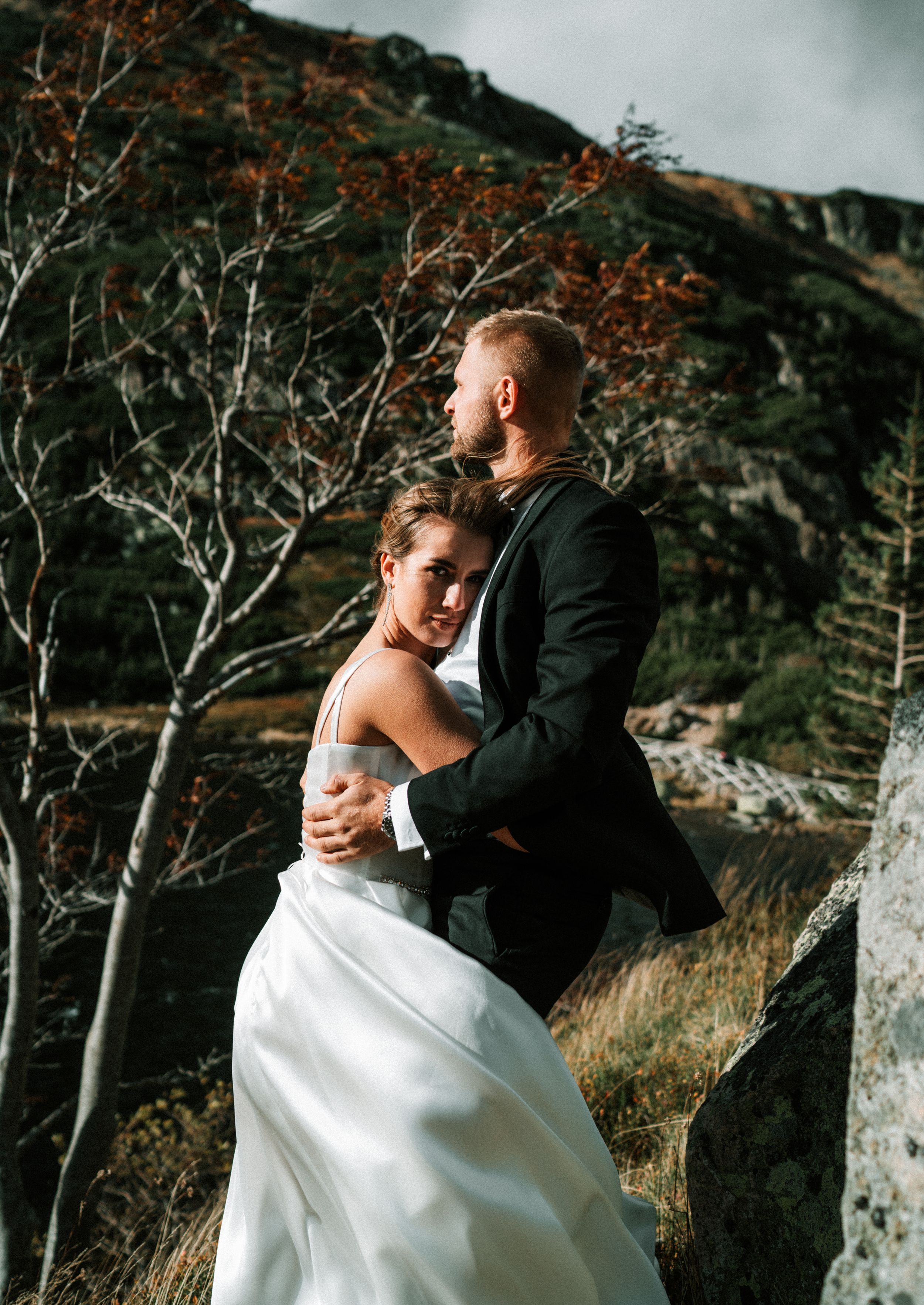 Wedding couple embraces against dramatic mountain backdrop with bare trees in autumn setting.