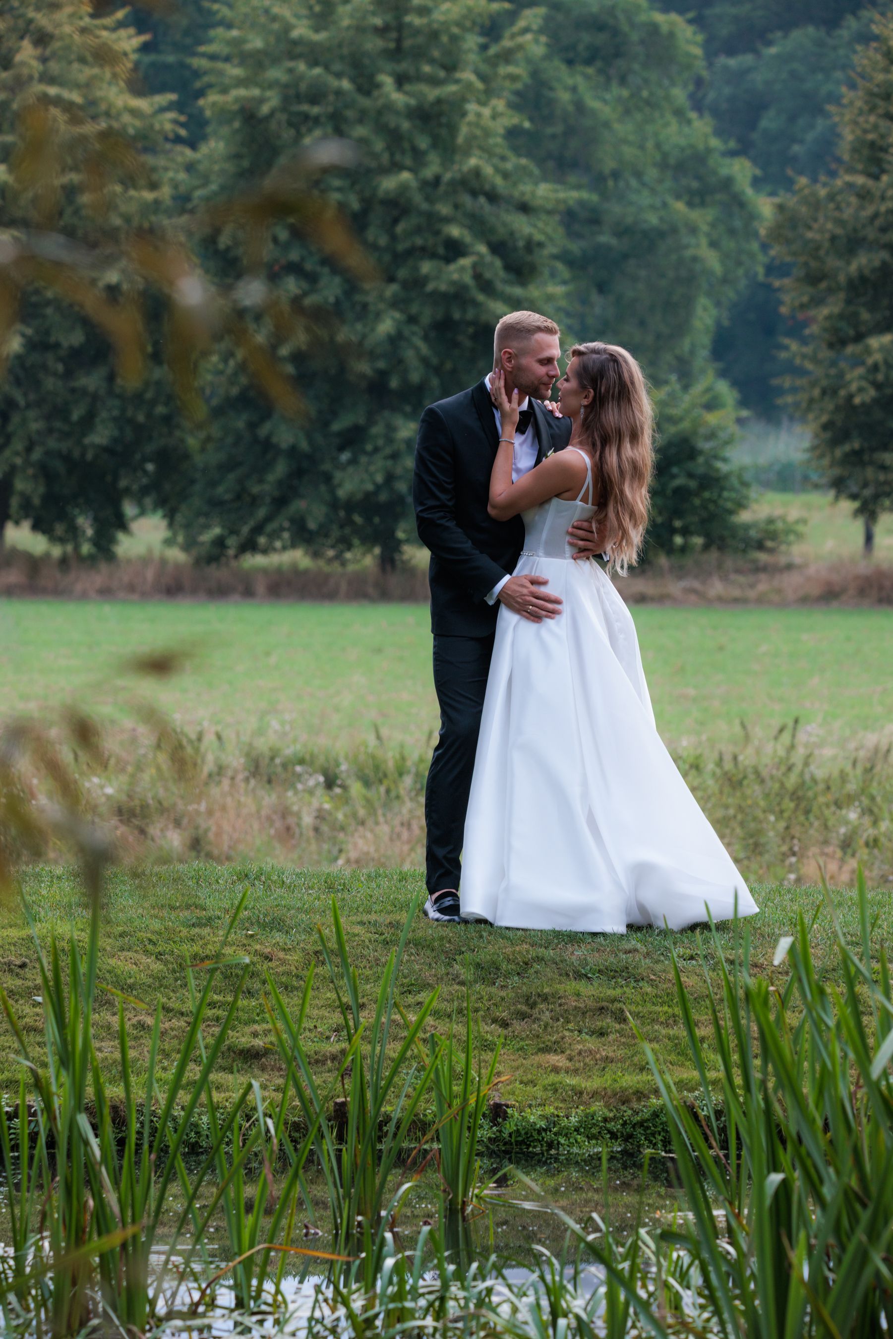 Romantic wedding couple embracing by a pond with trees in the background.