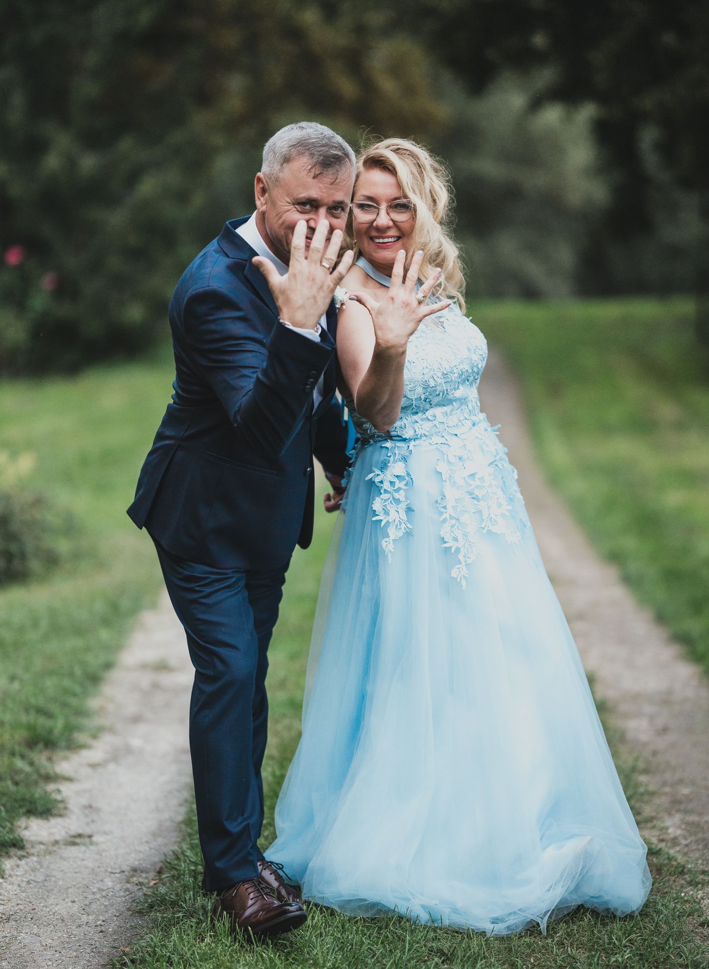 Couple sharing joyful moment showing off rings on a country path, bride wearing ethereal light blue gown.