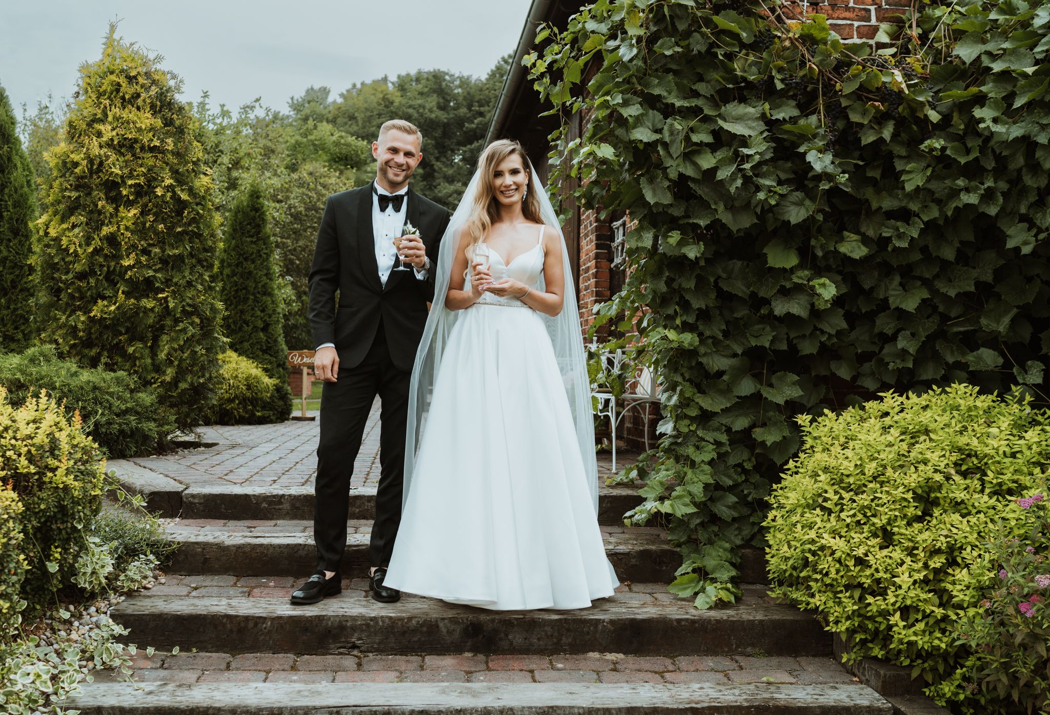 Wedding couple poses on garden steps surrounded by manicured hedges and greenery.