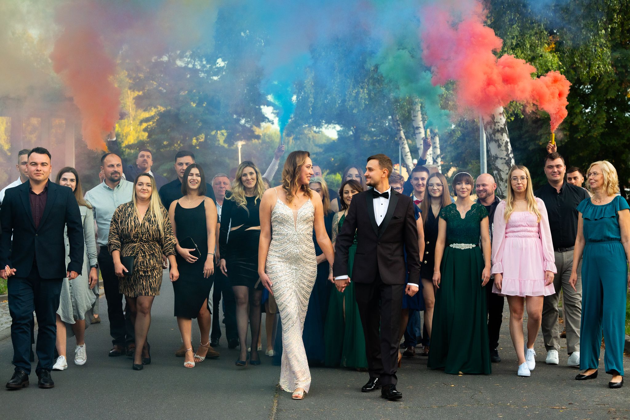Wedding party celebrating with colorful smoke bombs outdoors in formal attire.