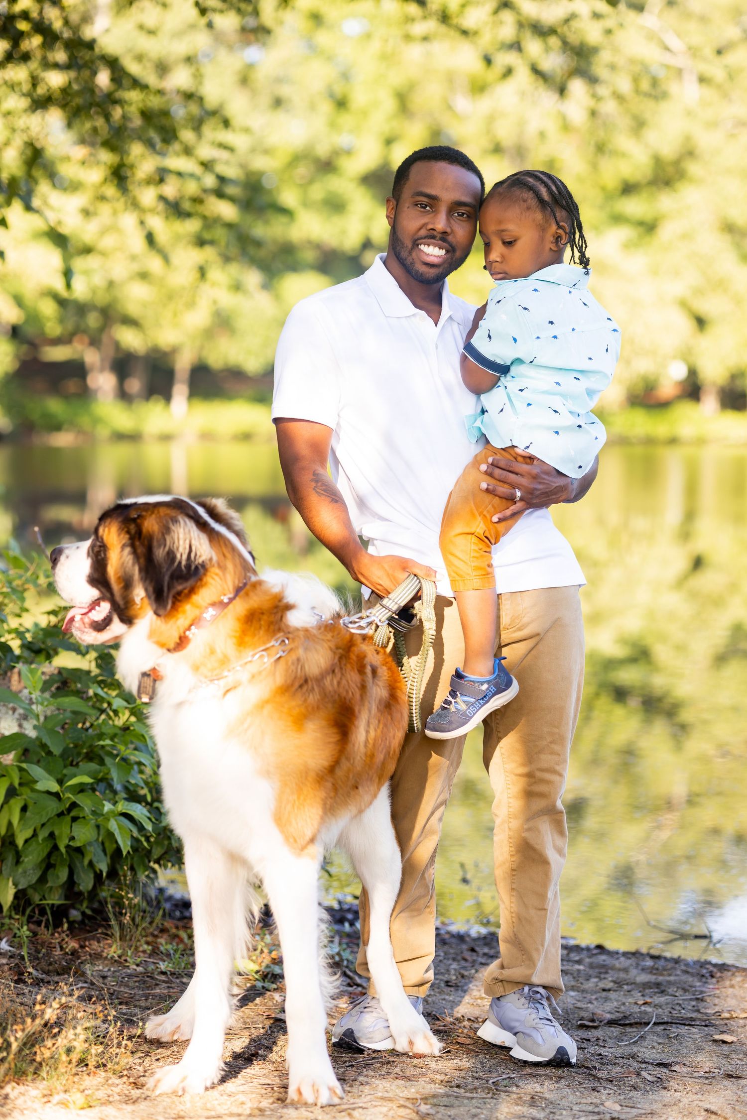 Parent holds child while standing with Saint Bernard dog by lakeside.