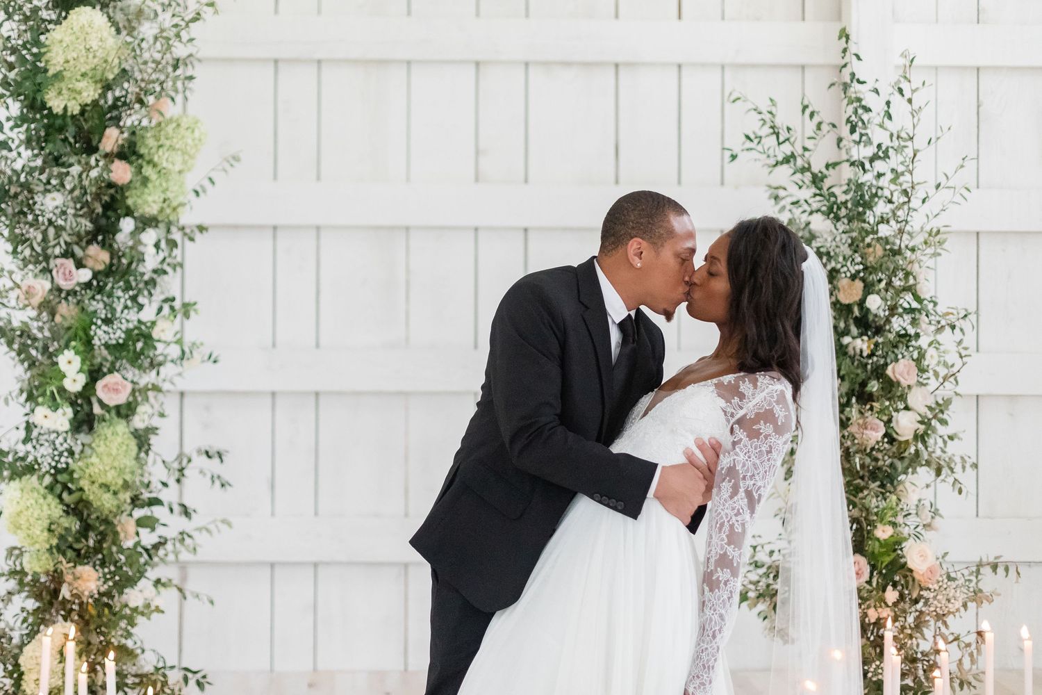 Elegant wedding ceremony setup with white floral arch and wooden chairs in a bright indoor venue.